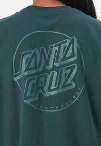 Grønn t-skjorte med en stor, lysegrønn "SANTA CRUZ" grafikklogo og en sirkulær ramme, laget av myk bomull.