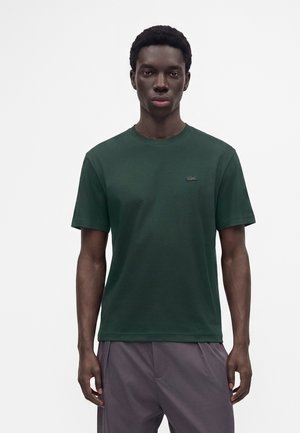 Ung mand med kort krøllet hår iført en mørkegrøn t-shirt med korte ærmer og grå bukser, stående foran en ensfarvet lys baggrund.