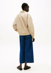 Giacca bomber beige con colletto a coste, vestibilità over, e polsini elastici, abbinata a pantaloni in denim scuri a gamba larga e scarpe nere slip-on.