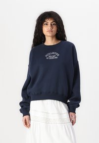 Temno modra majica s kapuco iz mehkega blaga, z rebrastim ovratnikom in vezenim logotipom "Hollister Sport & Leisure" spredaj.