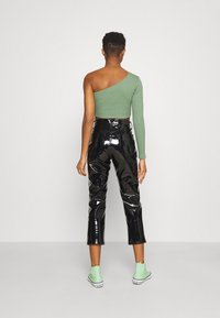 Person, der ein grünes Ein-Schulter-langärmliges Oberteil, glänzende schwarze cropped Pants und mintgrüne High-Top-Sneaker trägt, steht vor einem einfarbigen Hintergrund.