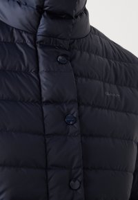 Navyblå dunjakke med vandret quiltning, der har en høj krave og skinnende knapper. "GANT" logo diskret trykt på stoffet.