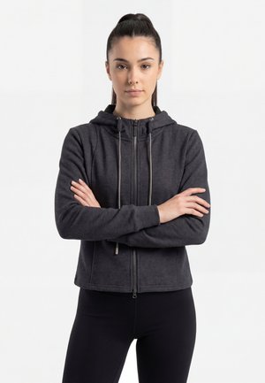 Nuori nainen, jolla on tummat hiukset poninhännällä, pukeutuneena mustaan vetoketjulliseen huppariin ja mustiin housuihin, seisoo käsivarret ristissä valkoista taustaa vasten.