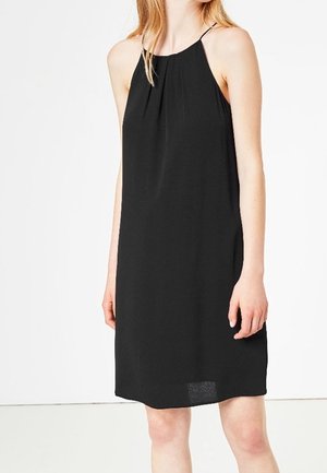 Vrouw draagt een mouwloze zwarte jurk tot op de knie met een hoge halslijn en dunne schouderbandjes, staand tegen een effen witte achtergrond.