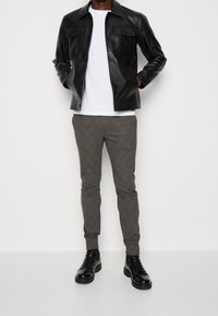 Homme portant une veste en cuir noir, un t-shirt blanc, un pantalon gris slim-fit et des bottes à lacets noires brillantes, se tenant les mains dans les poches.