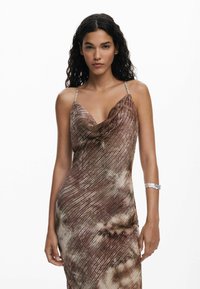 Barna mintás, ujjatlan ruha, galléros nyakkivágással és vékony pántokkal, lágyan áramló anyagtextúrával és finom fénylő hatással. Ezüst karpánt kiegészítő.