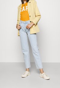 Blazer amarillo sobre camiseta naranja de GAP, jeans de tiro alto azul claro y zapatillas crema. Una pulsera de plata en la muñeca.