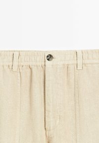 Un pantalon en lin beige présente une taille froncée et une fermeture à un bouton. Le tissu a une apparence légèrement texturée avec des plis frontaux.