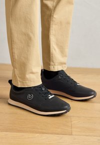Black athletic shoes with a perforated design, featuring laces and a light-colored sole. Worn with light beige pants on a wooden floor.