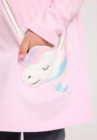 Maglietta rosa a maniche lunghe con una tasca di peluche bianca a forma di unicorno, dettagli della criniera nei toni pastello e riflessi di glitter, indossata su pantaloni scuri.