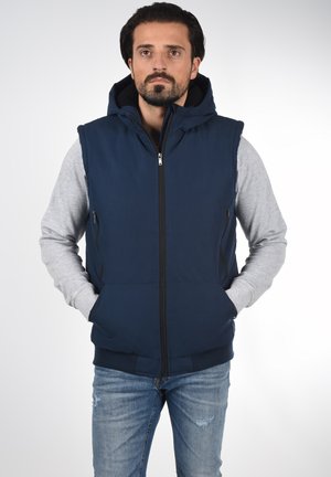 Navy blue sleeveless vest with a hood, synthetic fabric, front zipper, side pockets, layered over a gray long-sleeve shirt. Casual design.