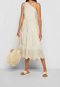 Vestido de un solo hombro, de color beige claro, con un diseño en capas, dobladillo de encaje de ojal y tejido fluido. Combinado con una bolsa tejida y sandalias tipo slider.