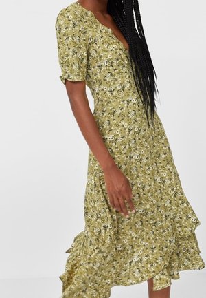 Femme aux longs cheveux tressés portant une robe midi fleurie vert clair avec manches courtes et ourlet à volants.