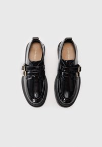Black patent leather shoes with a rounded toe, featuring dual buckle straps and a thick black sole. Textured surface, minimalistic design.