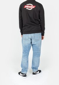 Sweat-shirt noir avec logo "Dickies" rouge dans le dos, associé à un jean bleu clair et des baskets noires avec des accents blancs.