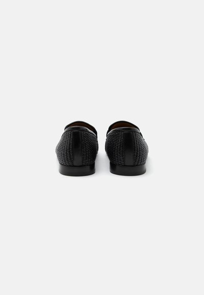 Μαύρα υφαντά δερμάτινα παπούτσια τύπου slip-on, εμφανιζόμενα από πίσω σε λευκό φόντο, που αναδεικνύουν την υφή και τα επίπεδα πέλματα.