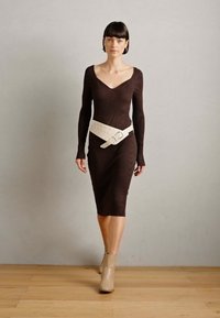 Vestido largo de manga larga ribeteado en marrón con un escote en V profundo y un cinturón ancho blanco. La modelo lleva botas de tobillo beige, de pie sobre un suelo de madera.