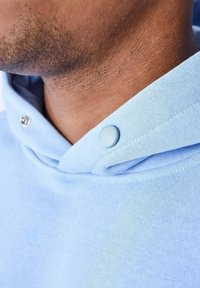 Sudadera azul claro hecha de una tela suave, con cuello cosido, cordón ajustable y dos broches visibles de color azul claro cerca del collar.
