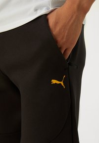 Sorte joggerbukser lavet af blødt stof, med et gult Puma-logo, lommer og en snøre detalje i taljen.