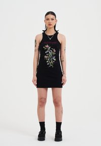 Schwarzes ärmelloses Rippkleid mit buntem Blumenstickerei und Grafikdesign. Das Model trägt schwarze Plateauschuhe und Socken.