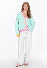 Pastellfarbener Cardigan mit Herzmustern, mit einem pinken Knopfstreifen, oversize Passform und gerippten Bündchen. Getragen über hellblauen Jeans.