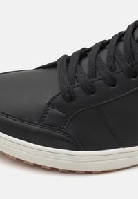 Black leather sneaker with textured finish, featuring black laces, white midsole, and a patterned rubber outsole. Close-up view highlights stitching details.