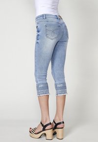 Femme portant un jean court bleu clair avec des poignets brodés et des sandales compensées à brides noires, debout sur un fond uni.