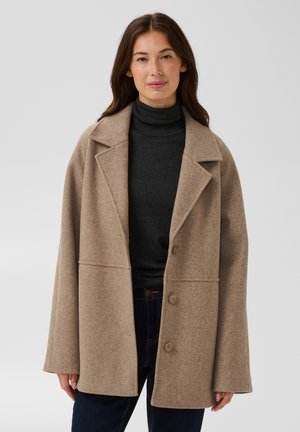 Femme portant un manteau en laine beige oversize, un pull col roulé noir et un pantalon foncé sur un fond uni gris clair.