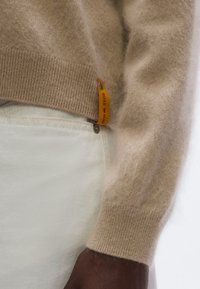 Beżowy sweter dziany o miękkiej fakturze, z prążkowanymi mankietami i kontrastową metką z logo w kolorach pomarańczowym i czerwonym przyszytą do dołu.