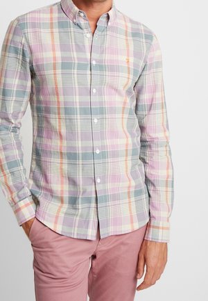 Mann trägt pastell-karierte Hemd mit Knöpfen und rosa Hose, steht mit einer Hand in der Tasche vor einfarbigem Hintergrund.