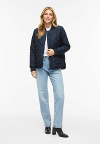 Prešita mornarsko modra bomber jakna z rebrastimi manšetami, sprednjim zadrgo in stranskimi žepi, kombinirana z svetlo moderimi kavbojkami z ravnimi hlačnicami in črnimi petkami.