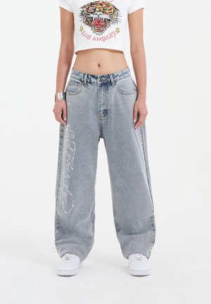 Jeans baggy azzurri chiari con una finitura sbiadita, tasche ampie e un intricato ricamo bianco lungo il lato sinistro, indossati con scarpe da ginnastica bianche.
