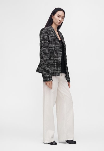 Femme debout portant un blazer à carreaux noir et blanc, un haut noir, un pantalon blanc large et des chaussures noires sur un fond uni.