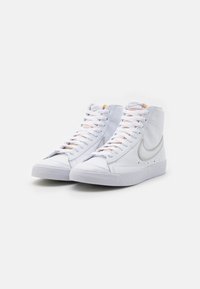 Vysoké bílé tenisky vyrobené z hladké syntetické kůže s texturovanou gumovou podrážkou, zdobené světlými tkaničkami a kovovým logem ve tvaru klíče.