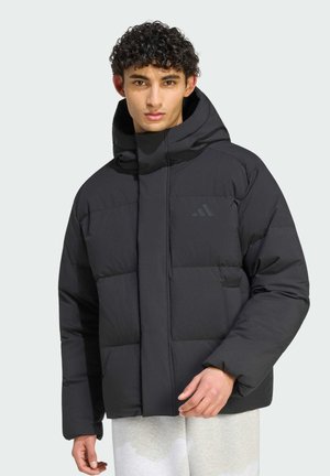 Giovane uomo che indossa una giacca imbottita nera con cappuccio e pantaloni chiari, in piedi davanti a uno sfondo chiaro e uniforme.