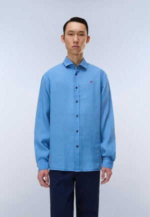 Młody mężczyzna stojący, ubrany w jasnoniebieską, zapinaną na guziki koszulę z długim rękawem oraz ciemne spodnie na jednolitym, jasnym tle.