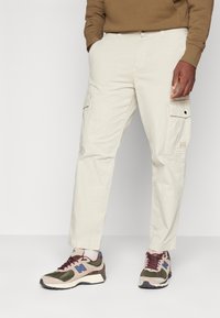 Béžové cargo kalhoty mají dvě boční kapsy, rovný střih nohavic a prošití. Kombinovány s vícebarevnými teniskami.