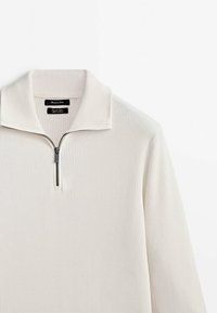 Pull-over en crème clair avec une texture côtelée, doté d'un col montant et d'une fermeture à mi-glissière. Design minimaliste.