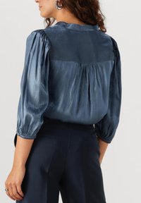 Satijnen marineblauwe blouse met opgezette mouwen, geplooid detail aan de achterkant en een soepele textuur. Gepaard met op maat gemaakte donkere broek.