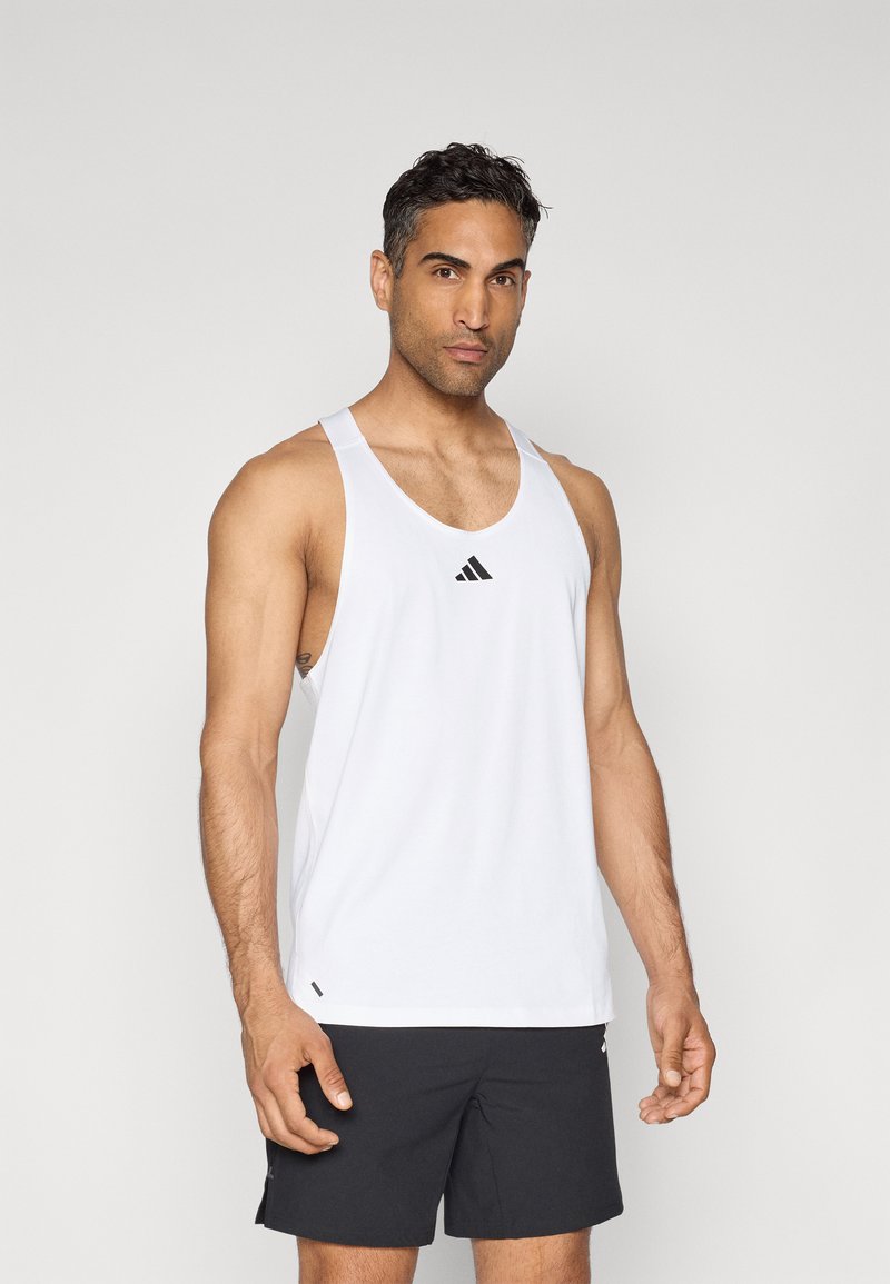 Débardeur de sport blanc sans manches avec un décolleté arrondi et un logo Adidas noir sur la poitrine, associé à un short noir. Matière lisse.