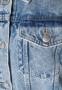 Světle modrá džínová bunda s kovovými stříbrnými doplňky, kontrastním prošíváním a dvěma náprsními kapsami s klopami. Celkově oprýskaná textura.