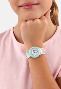 Eine Kinderuhr mit einem mintgrünen Kunststoffgehäuse und einem Stoffarmband mit floralem Muster. Das Ziffernblatt hat klare Stundenmarkierungen und Zeiger.
