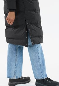 Schwarze Steppjacke mit Druckknöpfen, getragen über hellblauen weiten Jeans und schwarzen Stiefeln. Die Jacke weist eine glatte, isolierte Textur auf.