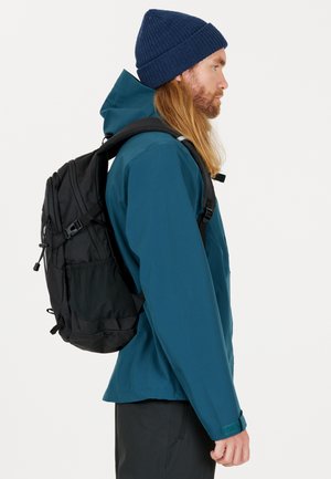 Teal vattentät jacka, svart ryggsäck och blå stickad mössa. Sidoprofilen visar långt hår, textil med struktur och justerbara remmar.
