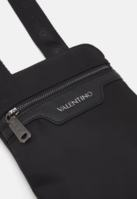 Černá nylonová taška s uzávěrem na zip, texturovaným předním panelem a názvem značky "VALENTINO" vytištěným stříbrně na horní části. Součástí jsou nastavitelné popruhy.