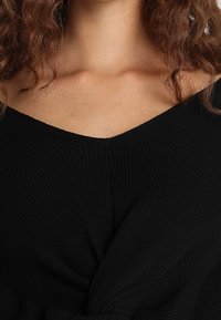 Svart stickad topp med V-ringning och vridet framstycke. Tyget har en ribbad yta med en jämn finish.