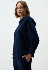 Donkerblauwe hoodie van zacht, textuurachtig materiaal met verlaagde schouders en een relaxte pasvorm. Voorzien van een voorzak en lange mouwen.
