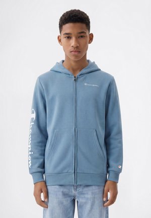 Teenage boy wearing a blue zip-up hoodie with front pockets and Champion branding on chest, sleeve, and cuff, standing against a plain background.