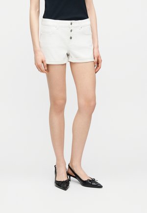 Kobieta w białych podwiniętych krótkich spodenkach dżinsowych z trzema guzikami z przodu, czarnej bluzce i czarnych sandałkach na niskim obcasie z kokardkami.