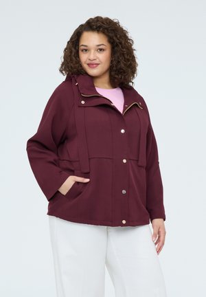 Kobieta w bordowej kurtce na zatrzaski z kapturem, w białych spodniach, stojąca z jedną ręką w kieszeni.